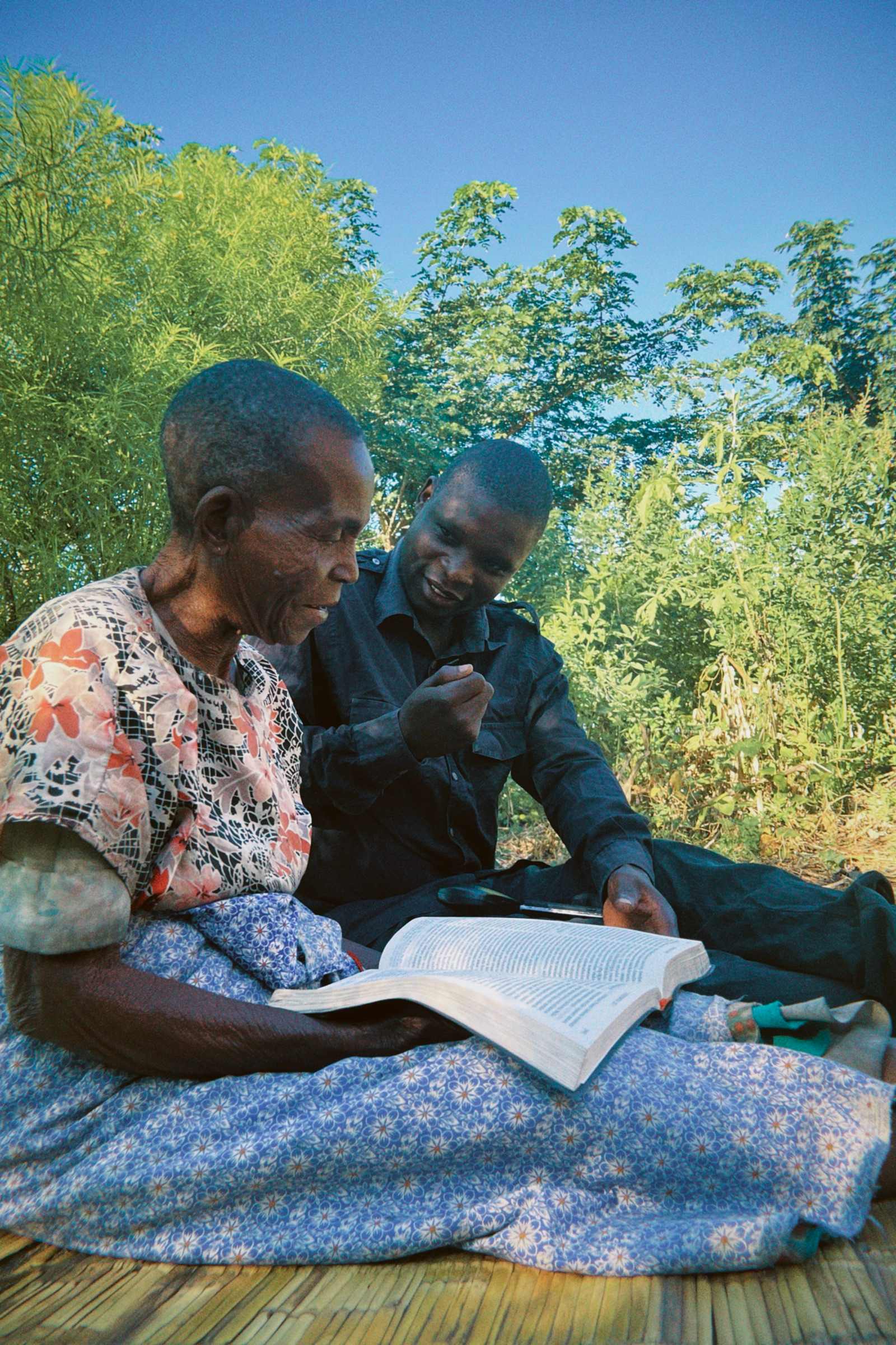 A grandmother and a man read the Bible together, sitting on a mat outdoors