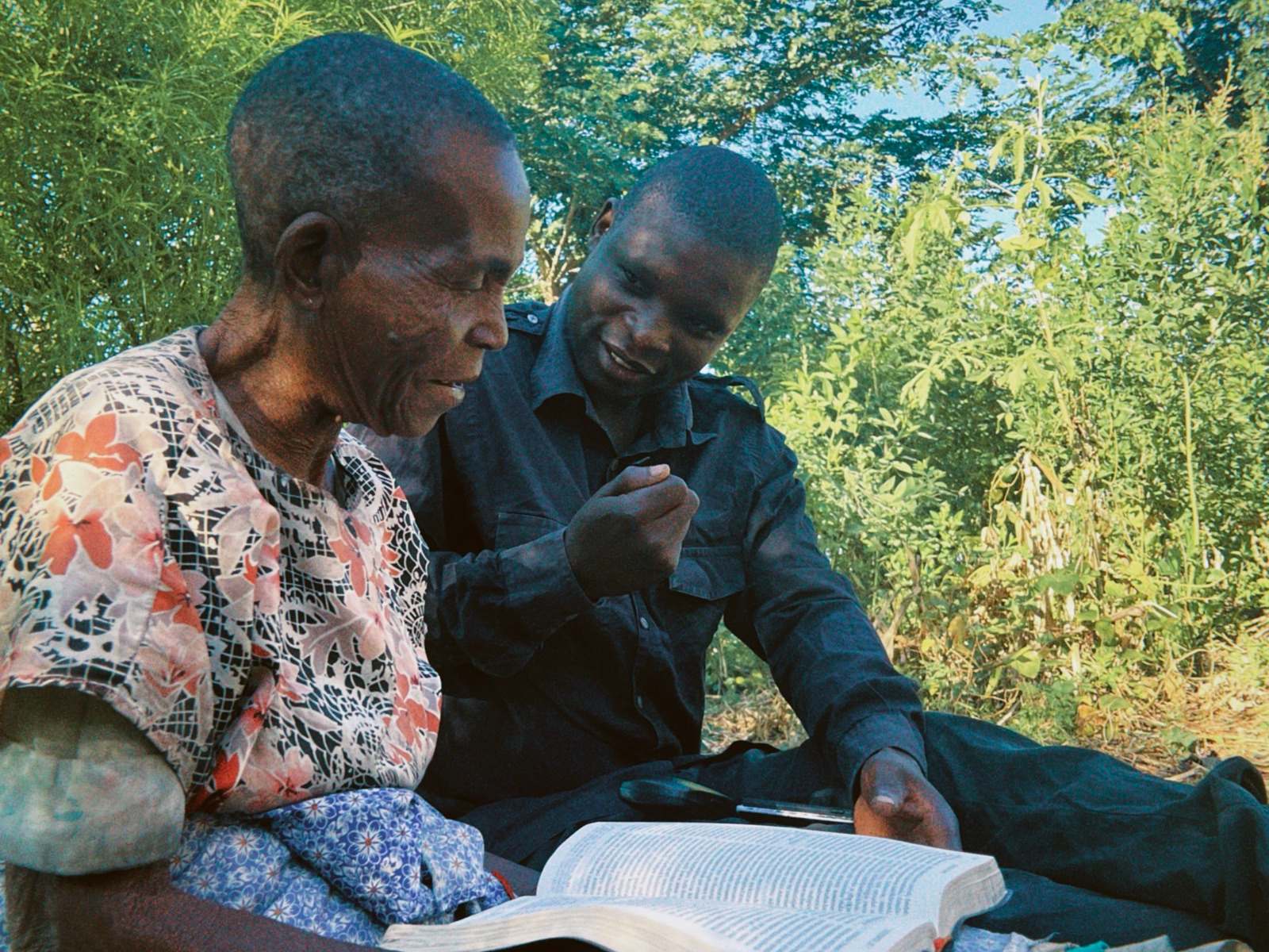 A grandmother and a man read the Bible together, sitting on a mat outdoors